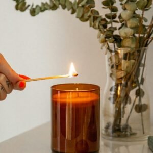 Bougie dans un pot en verre ambré, allumée à l’allumette, devant un bouquet d’eucalyptus